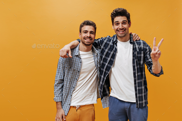 Cheerful young men in plaid blue shirts, white t-shirts and colorful pants  pose on orange backgroun Stock Photo by look_studio