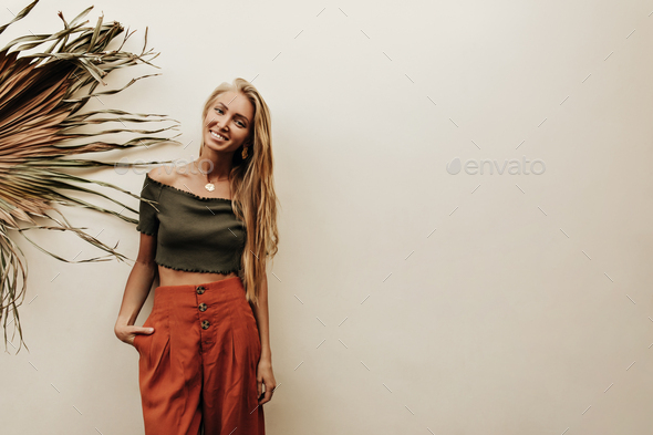 Attractive blonde long-haired happy woman in cropped khaki top and red loose pants smiles and poses