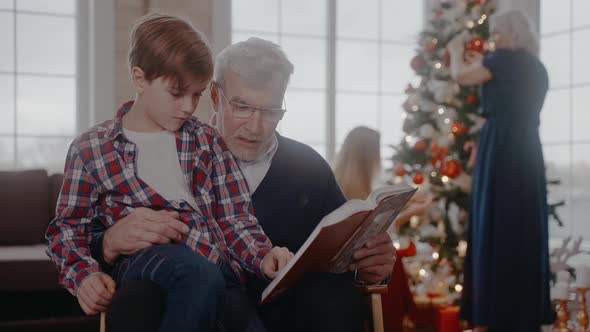 Grandad Reading a Book with Grandson Sitting on a Chair at Christmas Celebration alt