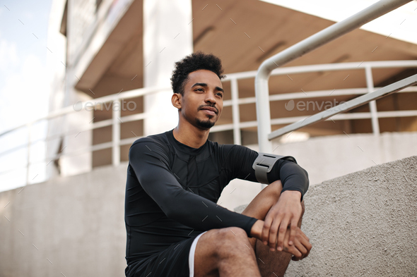 Portrait of young curly bearded dark-skinned man in black long-sleeved t-shirt and shorts sitting o