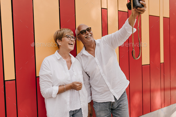 Cheerful woman with blonde hair in white blouse laughing and making photo with man with mustache in