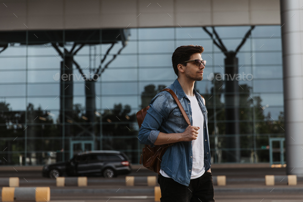 Free Photo  Handsome man in denim jacket and white tshirt poses outside  portrait of brunette guy in sunglasses holding bag