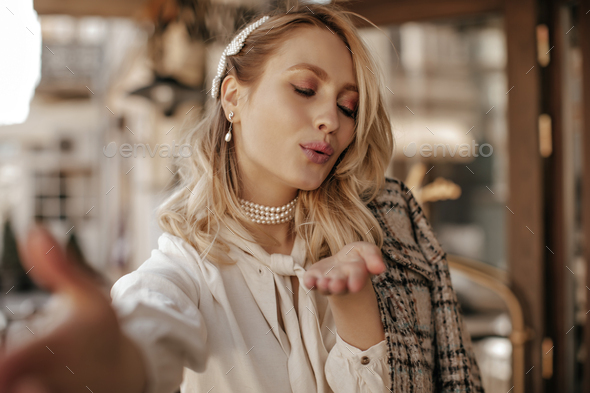 Attractive young blonde curly woman in white blouse and checkered coat blows kiss and takes selfie