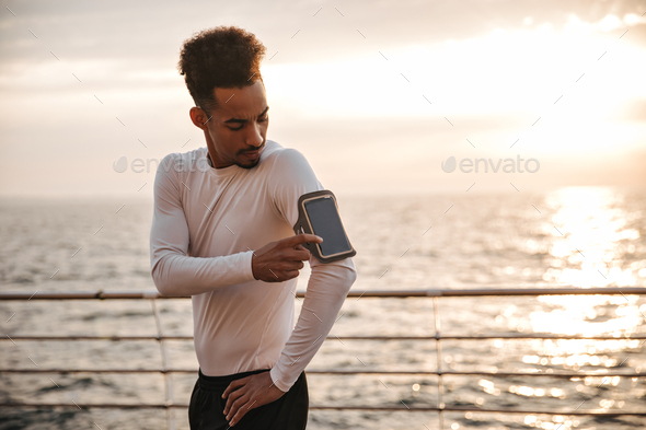 Curly dark-skinned cool man in white long-sleeved t-shirt and black shorts taps on phone screen and