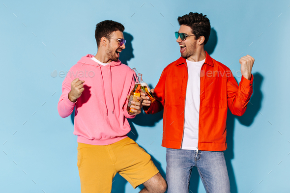Good-humored happy friends in colorful outfits and sunglasses have fun and clink beer bottles on bl