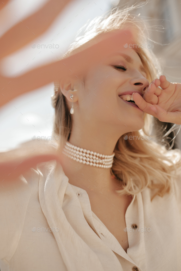 Cheerful blonde curly woman in pearl necklace and white blouse giggles and poses outside. Attractiv