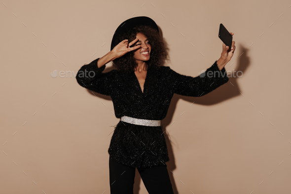 Modern lady with wavy hair in stylish black clothes and cool hat showing peace sign and making self