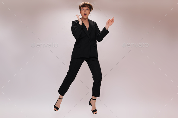 Pretty lady in suit jumps on isolated background. Cheerful short-haired woman in black jacket and p