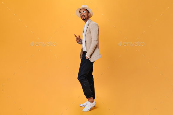 Guy in hat and suit posing on isolated background. Portrait of dark-skinned man in black pants, whi