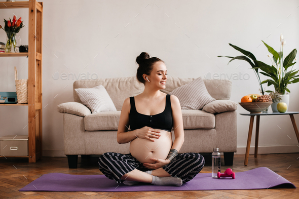 Joyful brunette pregnant woman in cropped top and black pants smiles, listening to music in white w