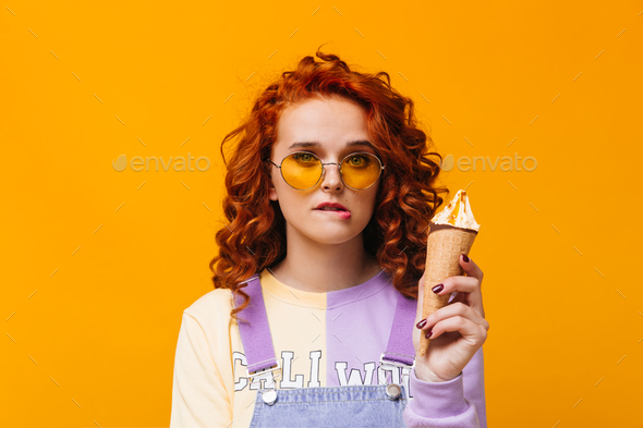 Blue-eyed girl with curls bites her lip, looks into camera and holds ice cream in waffle