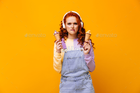 Redhead woman in headphones bites her lip, looks into camera and keeps ice cream in waffle