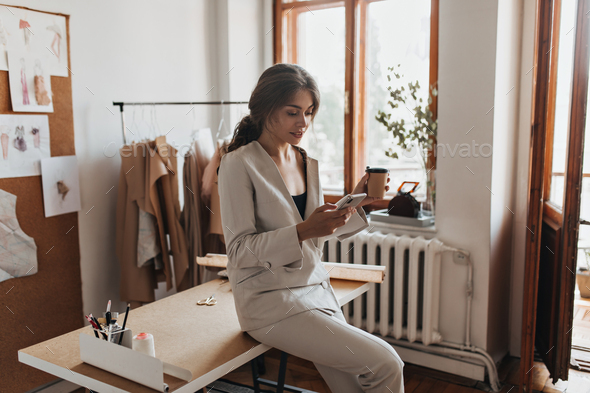 Dark haired lady in white suit holding coffee and phone. Charming woman in linen pants and beige bl