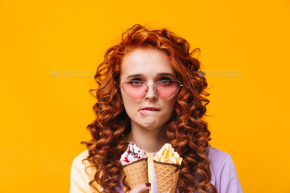 Blue-eyed girl in pink glasses bites her lip and looks into camera. Woman holding ice cream
