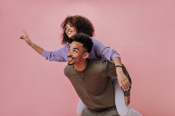 Curly girl jumped on her boyfriends back and showing peace sign. Dark-skinned couple has fun and sm - Stock Photo - Images
