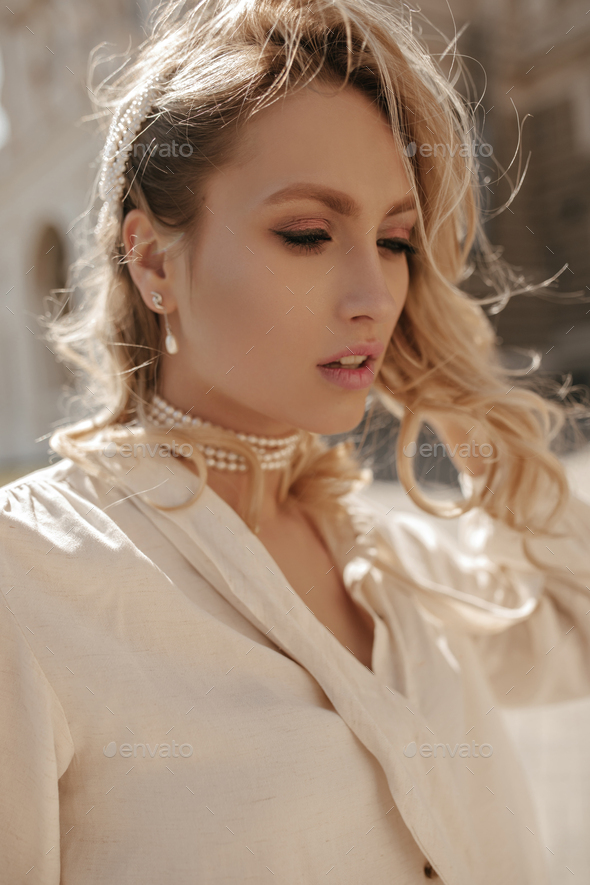 Attractive pretty blonde woman in white blouse and pearl necklace looks down and walls outside.