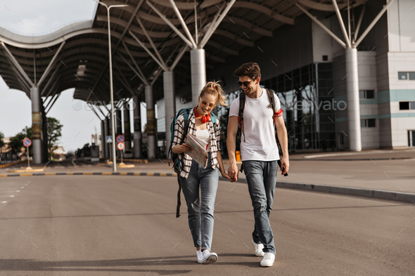 Cool blonde woman in jeans, plaid shirt and brunette guy in white tee-shirt walks along roadside in