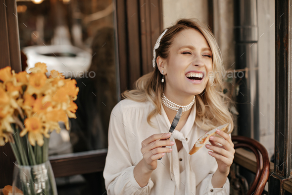 Cheerful happy blonde woman in white blouse laughs sincerely, holds knife and peace of bread and re
