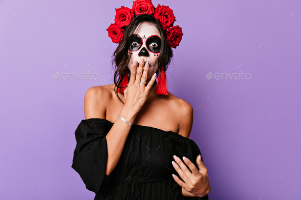 Shocked beautiful woman with face painting covering her mouth on purple background. Stylish latin l