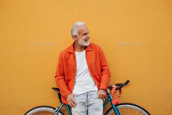 Smiling man in white shirt poses with bicycle on orange background. Gray-haired stylish guy in brig