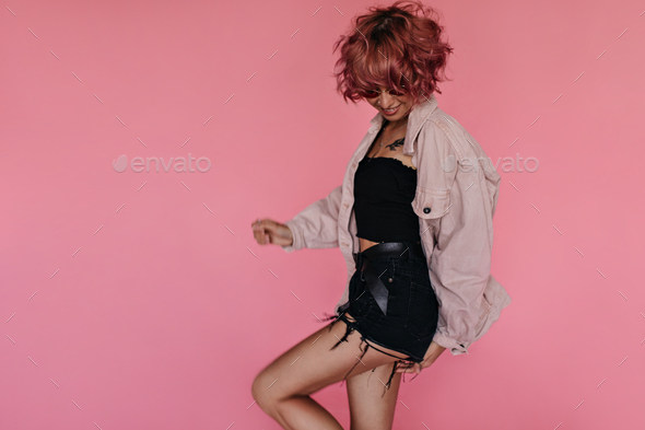 Curly pink-haired woman in black denim shorts and cropped top moves on isolated. Happy girl in over