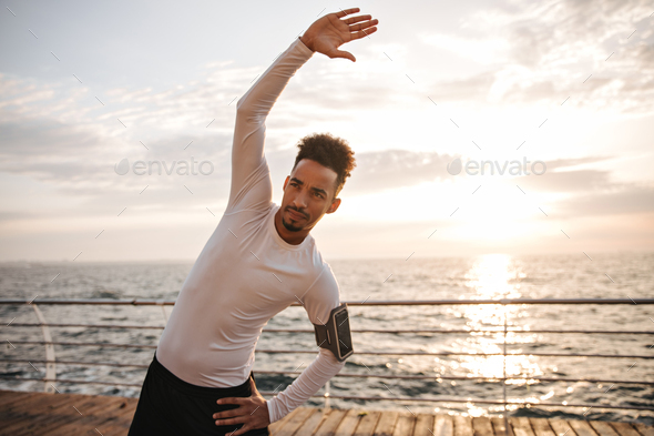 Curly young dark-skinned man in white long-sleeved t-shirt and black shorts stretches, rises arms a