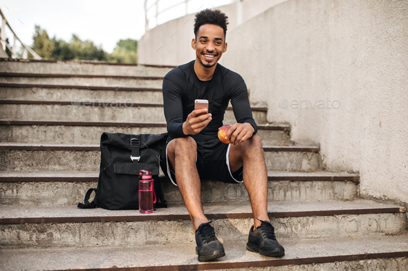 Cool young curly bearded dark-skinned man in black shorts and long-sleeved t-shirt smiles, sits on