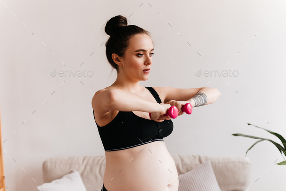 Young pregnant woman in black cropped top holds pink little dumbbells. Brunette girl doing sport in