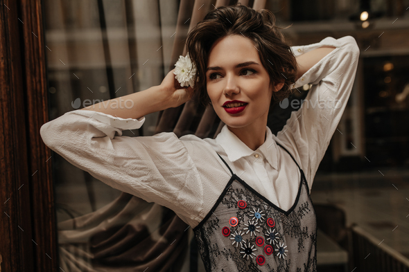Beautiful lady with curly hair and red lips looking away inside. Cool brunette woman in light blous