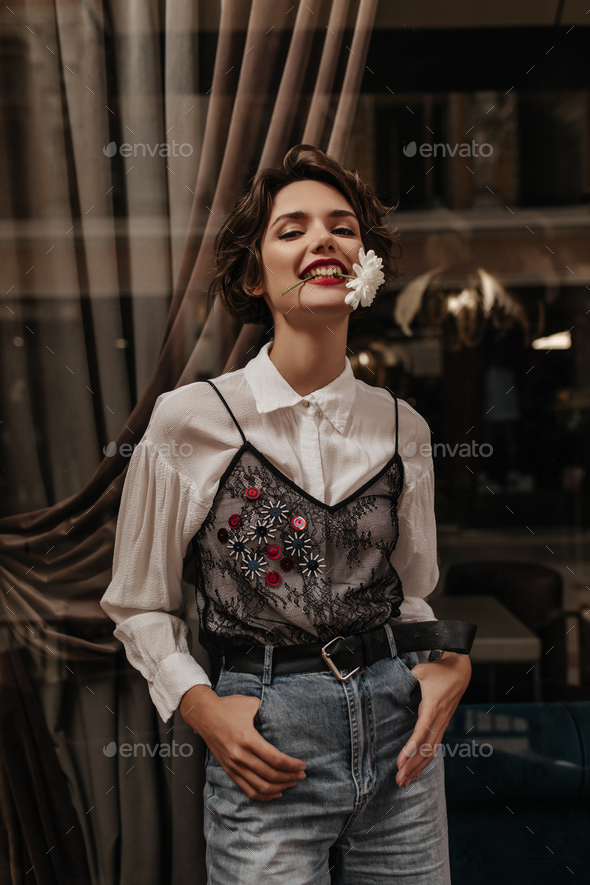 Positive woman with brunette hair in blouse with lace and jeans poses with flower in mouth inside.