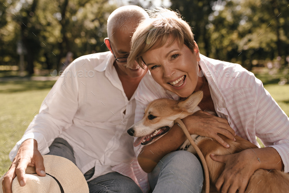Happy lady with short hairstyle in pink shirt laughing, hugging dog and posing with grey haired man
