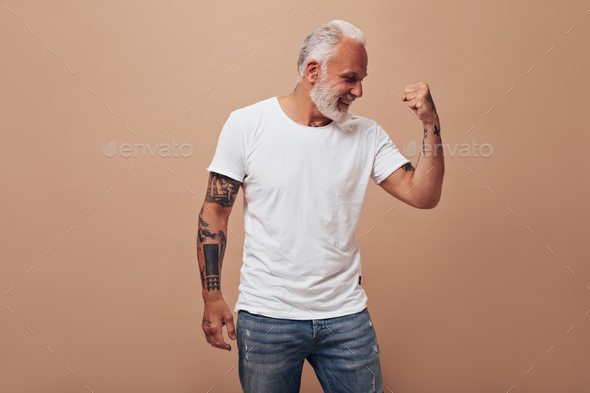Charming man in jeans and white shirt demonstrates biceps. Attractive guy in bright clothes in tatt