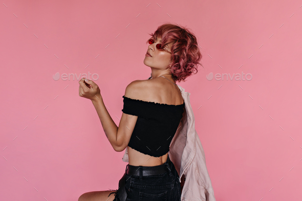 Pretty curly woman in cropped top and black denim shorts poses on pink background. Cute stylish tat