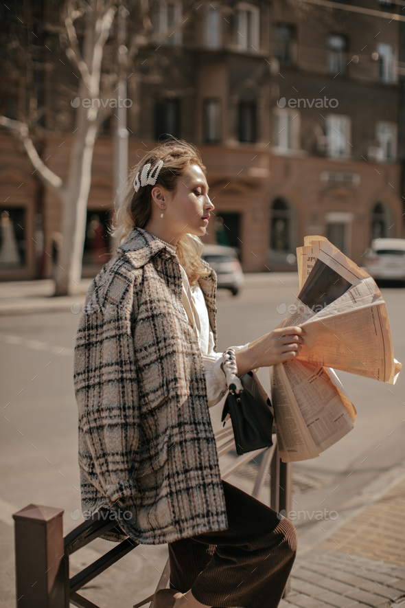Calm blonde woman in velvet brown pants, tweed checkered coat poses outside and reads newspaper in
