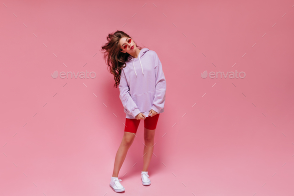 Long-haired girl in cycling shorts and purple oversized hoodie poses on pink background. Portrait o