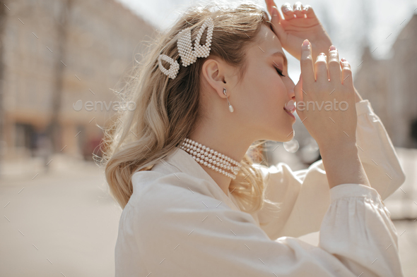 Calm young attractive blonde curly woman in pearl jewelry poses outside. Charming beautiful girl in