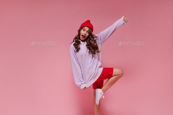 Attractive woman in red hat, cycling shorts and purple hoodie moves on pink background. Curly girl