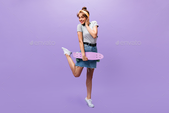 Smiling lady in denim outfit poses with longboard. Funny young woman in grey shirt and trendy skirt