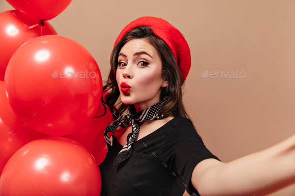 Curly girl in red beret and silk scarf whistles, holds balloons and makes selfie