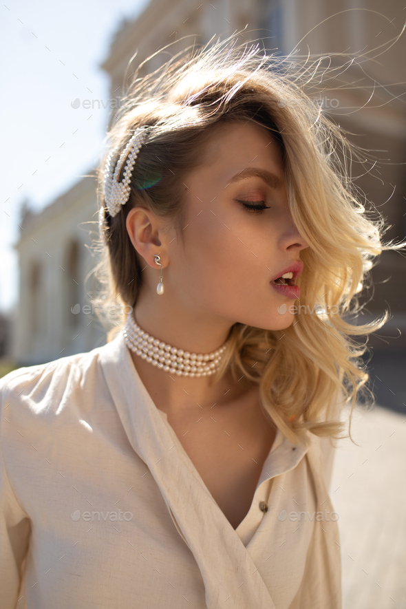 Young beautiful curly blonde woman in beige blouse and pearl necklace poses with closed eyes and wa