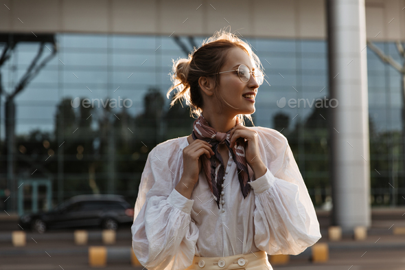 Blonde woman touches her silk brown scarf. Attractive young girl in eyeglasses and white blouse smi