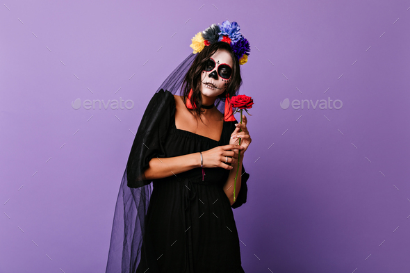 Upset latin girl with flowers on black veil posing in halloween. Brunette lady with zombie face pai