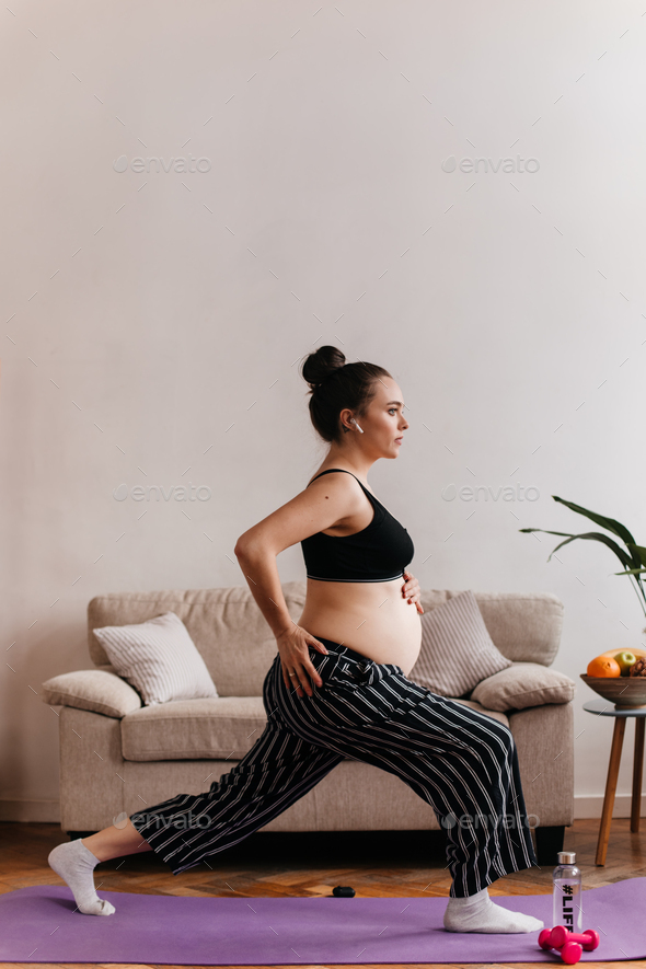 Healthy pregnant woman in black loose pants and cropped top stretches in living room. Brunette girl