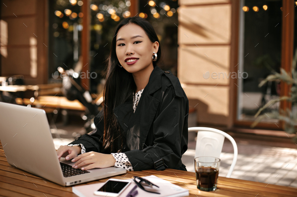Portrait of brunette woman in black trench coat works outside with laptop. Tanned lady in good mood