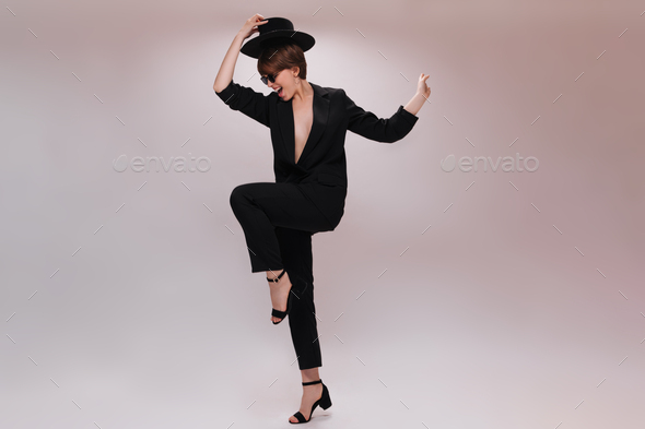 Joyful lady in suit takes off hat and jumps on white background. Pretty woman in black jacket and p