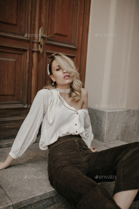 Grey-eyed blonde lady in velvet brown pants and stylish light blouse sits in relaxed pose on floor
