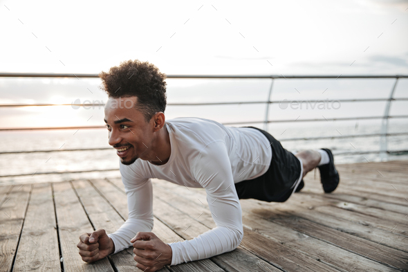 Happy excited active dark-skinned man in black shorts and white t-shirt smiles and does plank. Cool