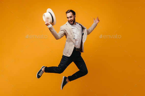 Stylish guy takes off hat and jumps high on isolated background.