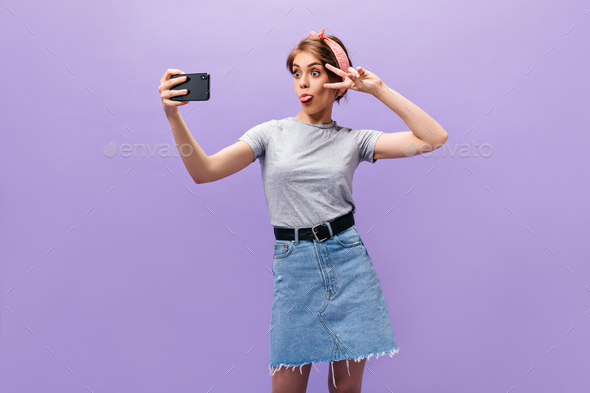 Girl in denim skirt demonstrates tongue, shows peace sign and takes selfie. Funny woman in good moo