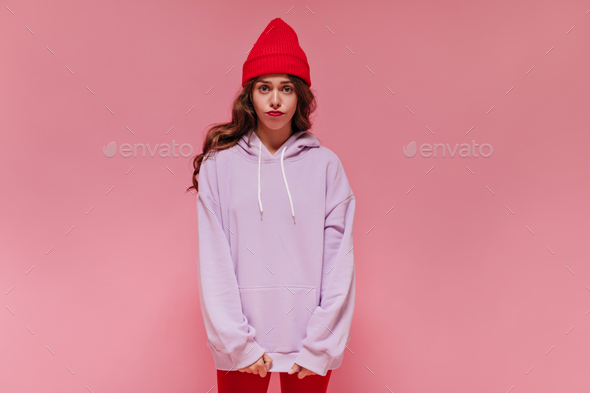 Sad woman in red hat, cycling shorts and purple hoodie poses on pink background. Brunette curly gir